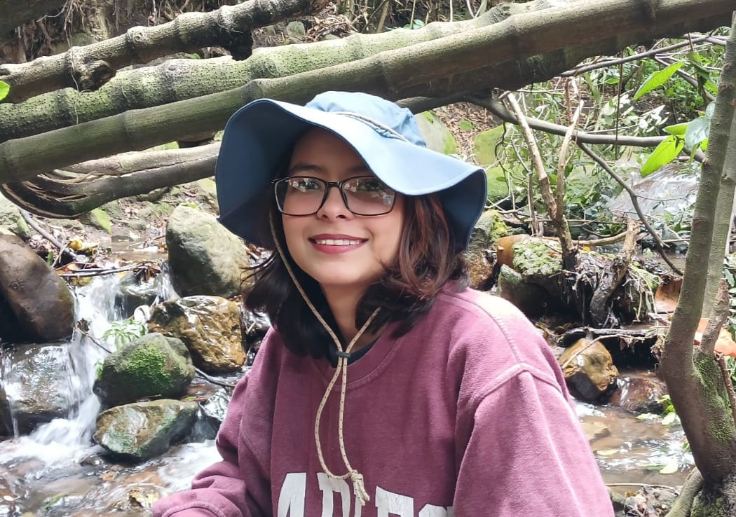 A person with long brown hair and glasses wearing outdoors clothing next to a rocky stream